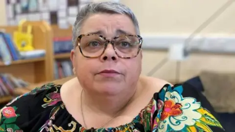 Maxine Thompson-Curl has grey hair and tortoiseshell glasses. She is looking serious and looking just away from the camera. She is wearing a colourful patterned dress.