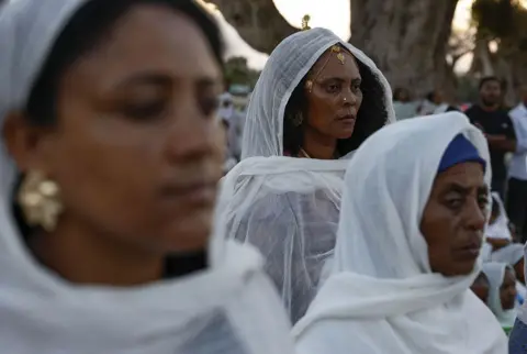 Anadolu via Getty Images Women in white cotton scarves and head coverings look on. 