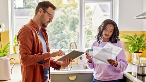 Getty Images A young couple stand in their kitchen looking at a tablet and some paperwork.