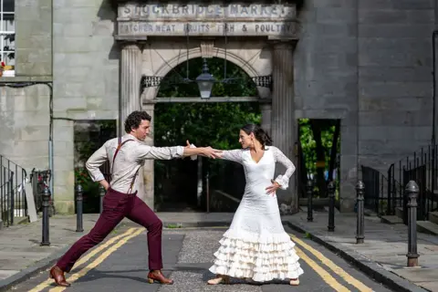 Roberto Ricciuti/Getty Images Gabriel Aragú Fernando and María Garcia Lola pose in front of the Stockbridge Market during a photocall for ‘Lola: A Flamenco Love Story’ He is wearing smart, burgundy trousers and she is in a beautiful white Flamenco dress, her hair in a pony tail. They are both looking at each other in a dance pose intensely.