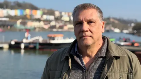 Man stood on Bristol Harbour with the water and colourful houses in the background. he wears a coat and a fleece