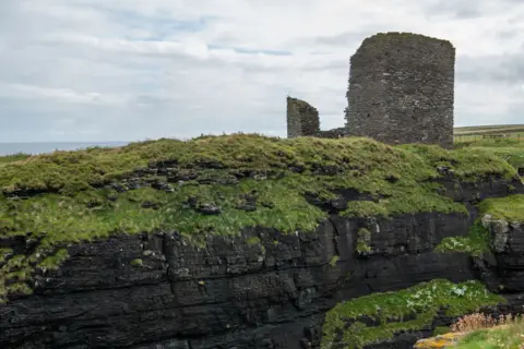 Historic Enviornment Scotland A dark grey stack of rocks forming a ruined castle sit upon a green grass bank with white clouds and a grey and blue sky in the background. 