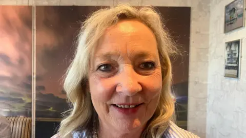 A woman with long blonde hair smiles at the camera as she wears a blue and white striped shirt. She is standing in front of a wall with a painting on it.