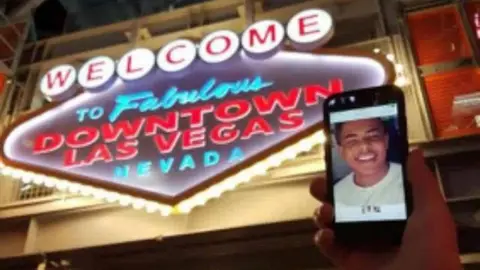 Family Breon pictured in Downtown Las Vegas. We see a hand holding an iPhone with a picture of him. The landmark is in the background. It is a dark evening and the sign is bright.