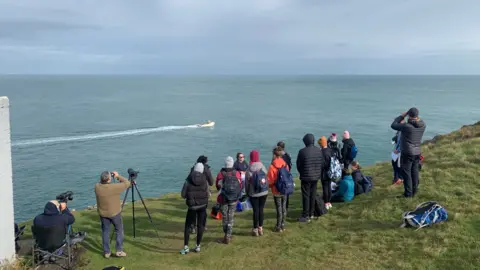 Sea Trust Wales A picture of a group of volunteers completing land-based surveys on the North Pembrokeshire coast. There are about 20 people observing the porpoises from the cliff. Some of them have cameras and binoculars. There is a boat in the sea and it is a sunny day. 