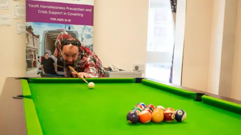 Bardsley Youth Project Simon is playing pool. He is holding the stick and is about to the hit the white ball and break up the other balls that are aligned in a triangle. He and is crouched down to do so. 