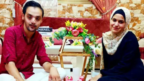 Mohamed and Noura sitting down in their home with some flowers between them. He is wearing a red shirt and she is wearing a dark blue dress and light patterned headscarf.
