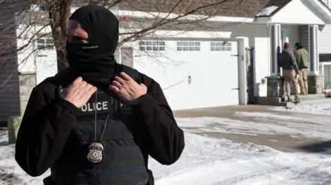 Reuters An ICE agent standing outside a white house. The agent has his face covered. 