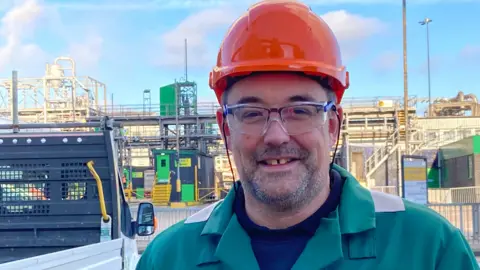 Jill Bennett/BBC Phil McNaughton standing outside at the sugar beet factory in Wissington. He is wearing green overall, protective eyewear and an orange hard hat.