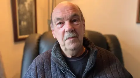Anton Hammer, 72 wearing a dark top with a balding head and dark eyes. He is sitting in an armchair and smiling into the camera.