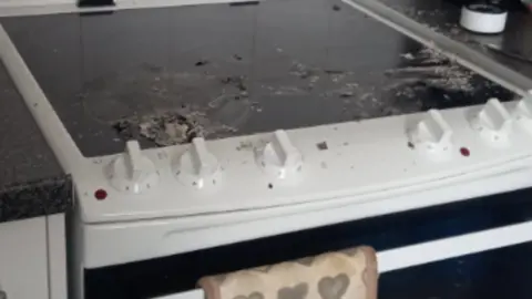 The picture shows a kitchen cooker with a black ceramic hob that appears to have been damaged by heat. There are burnt and melted remnants on the surface, suggesting something caught fire or was scorched. The hob has white control knobs at the front, and the oven door below has a heart-patterned oven glove hanging from the handle.