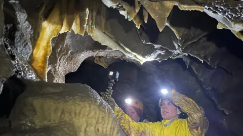 Two people in cave suits are examining yellowy orange cave features which are glistening with water. They are wearing helmets and lights and one of them is holding a small camera.