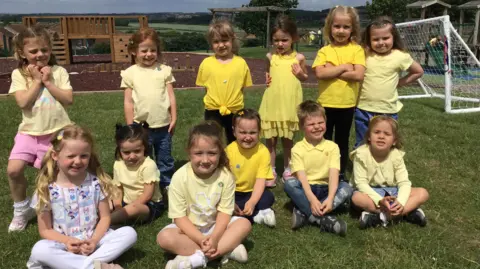 WINDHILL PRIMARY SCHOOL, MEXBOROUGH A group of young children on a school playing field wear yellow clothes or ribbons. They sit on the grass and pose for a photograph