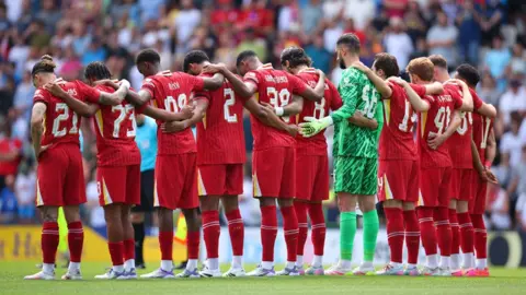Reuters Eleven Liverpool players, ten in the club's red kit and the goalkeeper wearing a green outfit, stand with their arms linked around their shoulders and their heads bowed as they observe a minute's silence on the pitch at Deepdale for their teammate Diogo Jota