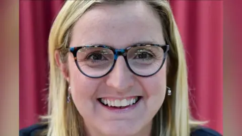 Labour Party Pamela Hargreaves is looking straight at the camera and smiling brightly. She has long blonde hair and blue/ brown glasses.