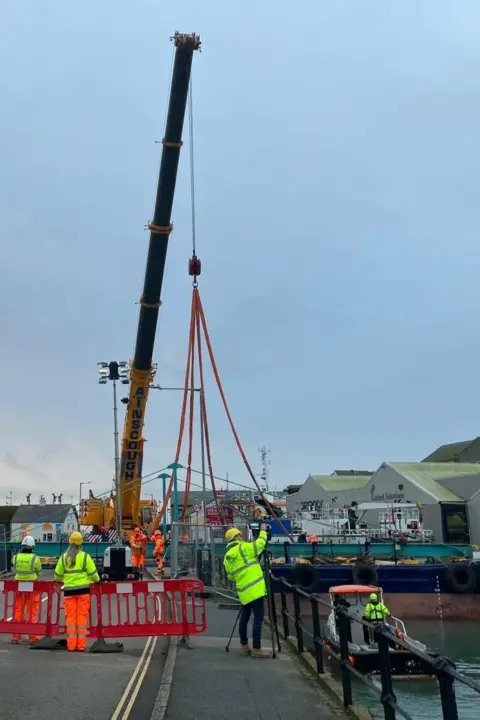 The crane is at full extension and its sling straps are around the bridge. In the foreground are people wearing helmets and high-viz jackets and trousers standing next to a safety barrier.