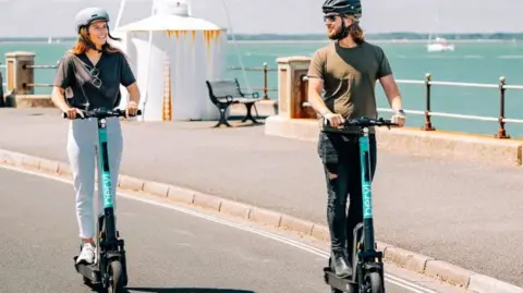 A man and a woman wearing helmets ride Beryl e‑scooters side by side along a sunny seafront road, with the sea and a small harbour structure visible in the background.