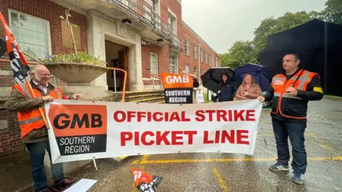 BBC Two people holding a GMB Union strike picket line banner on a rainy day outside a large red brick building.
