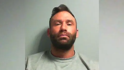 Mugshot of Luke Atkin, who has short, brown hair with a longer fringe. He has a brown beard and is wearing a grey sweatshirt. 