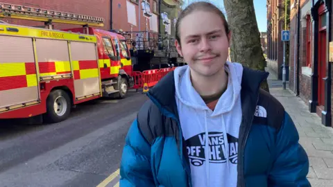 A man with tied-back hair and wearing a blue coat over a white Vans hooded top, stands on a pavement and smiles. Behind him there is a fire engine parked at the side of the road, with filming equipment in the background. 