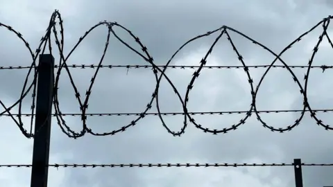 Getty Images Barbed wire against a grey, overcast sky. 