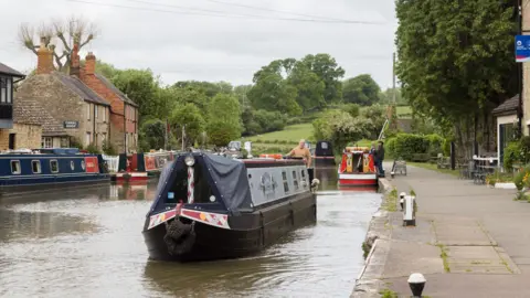 Canal & River Trust About five narrowboats on a canal, with a path on the right. There are buildings to the left and right, and trees behind the boats. A few people can be seen on the boats. 