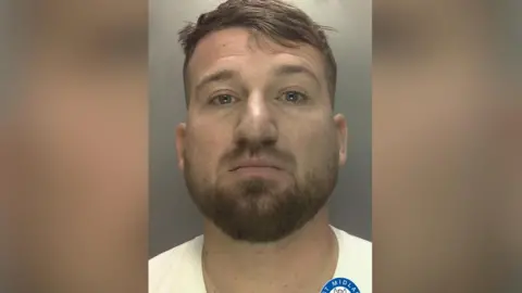 Dawid Dembler is pictured in a mugshot wearing a white t-shirt. He has light brown hair and a light brown beard.