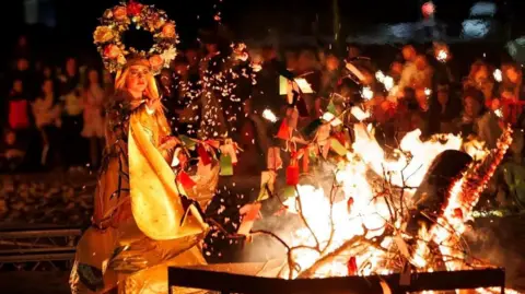 A woman wearing a dress decorated like a May Pole  casting items on a bright bonfire while crowds watch on.