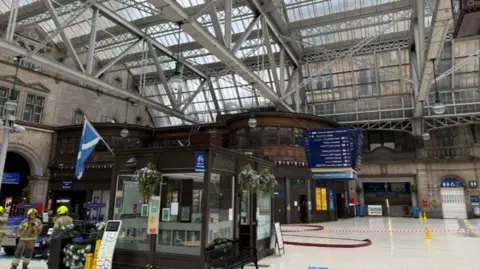 Network Rail two firefighters standing inside Glasgow's central station. part of the station is cordoned off an a large hose is visible.