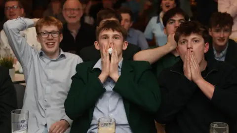 Liam McBurney/PA Wire Three young men are watching the golf. To the left of the picture, a man with ginger hair and glasses has his arm behind his head. In the middle is a man with a green jacket and striped shirt has his hand clasped nervously in front of his face. To the right, a young man wearing a black quarter-zip top is sitting with his hand clasped in front of his face watching the Masters golf intently and nervously. They are sitting in front of others who are watching nervously