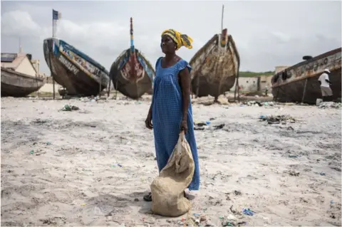 AFP A woman on a sandy beach with a sac standing in front of boats. She is wearing a blue dress and yellow head wrap and looking on.