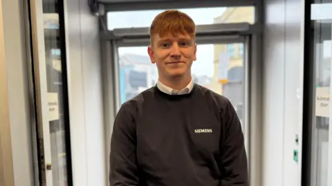 Robert Constantin/BBC A man with ginger hair in a grey jumper and white shirt smiles. 