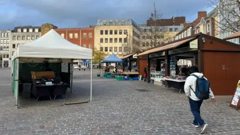 Alex Pope/BBC A man, with a white top on, blue trousers, with a blue backpack, walking through a town square, that has cobbles on the ground, a marque, tented stall to the left and back, and static stalls to the right. There are a number of buildings around the square, with trees in the distance. It also shows a blue sky. 