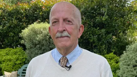 A man in his seventies with a moustache is stood in his garden on a sunny day.  He is smartly dressed, wearing a shirt, tie and v-neck jumper.  He is looking directly at the camera.