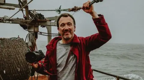 Pete Robinson Photographer Pete Robinson holding his camera and smiling on boat out at sea. He is wearing a red jacket over a grey t-shirt and a hat. The sea behind him is grey.