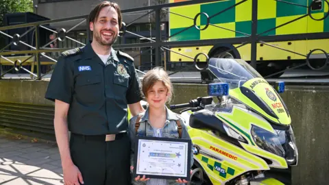 London Ambulance Service Emergency call handler Matthew Hawkins with Farah Alim-Palmer, who holds a certificate. They stand in front of a London Ambulance motorcycle at the emergency service's headquarters in Waterloo.