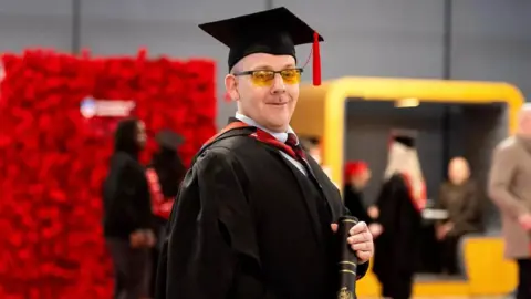 University of Lancashire Ben Constable in black graduation cap and gown holding his degree