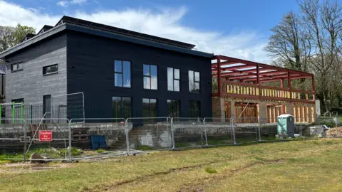 A picture of the outside of the building. There is a large grey building on the left of the screen with a number of windows and doors. There is a wooden structure to the right of the image, it is brown and golden in colour.