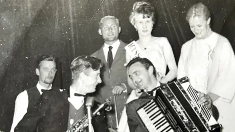 Doris Stout Six people standing together. One has an accordion and his bandmate has a saxophone. A third band member sits to the left of them but he has no instrument. Behind them stands Robert Shaw in a grey suit, white shirt, dark tie. A female winner in a light dress and a winner's sash, and Mary Ure in a white coat. They all appear to be enjoying themselves.