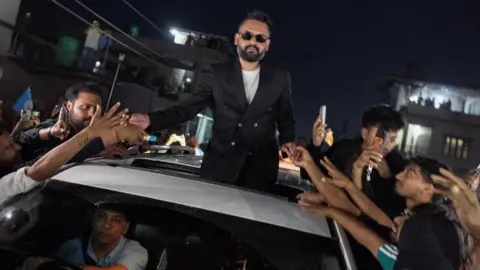 Balendra Shah standing out of a car's sunroof as he takes supporters' hands who are gathered around the car