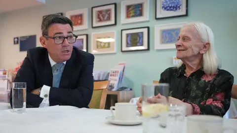 Andy Burnham sits at a table in a care home, talking face-to-face with a resident. The prime minister is in a dark suit and glasses and leans forward while speaking, and the other, with long white hair, sits opposite. Several glasses of water and cups are on the table, with framed artwork displayed on the wall behind them.