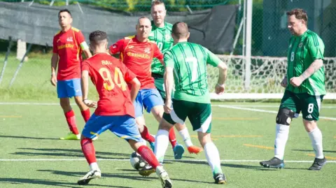 AICS Six male football players on a football pitch with a football. Three of the men on the left are wearing red shirts with blue shorts. To the right, three men are wearing green shirts and shorts. 