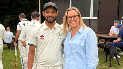 A smiling man dressed in cricket whites is stood next to a smiling woman in a blue shirt at a cricket ground