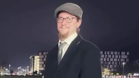 A man with a beard and glasses, he has long hair and wears a flat cap. He is dressed smartly in a coat, shirt and tie and stands in front of a city landscape at night. 