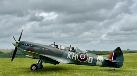 Monique A spitfire sits on the grass, preparing to take off with two people on board. The skies above are grey and full of clouds