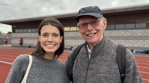 The photo shows a woman and a man smiling at the camera. The woman, on the left, has brown long hair and is wearing a grey jumper. The man, on the right, has a grey jacket on. He's also wearing glasses and a grey cap. They're standing on a brown athletics track. There is also a grey concrete building behind them. 