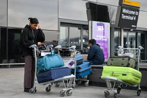 Getty Images A man and a women with suitcases piled on trolleys outside Terminal 4. The woman is looking at a phone and the man is sitting down on a wall