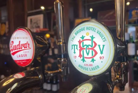 Two beer taps, one for Budvar and the other for the Bridge Hotel Vaults house lager. The badge is white with a green and red emblem.