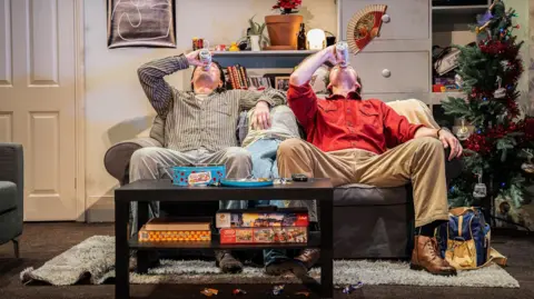 Barn Theatre Two men sit on a sofa in a living room with their heads tilted back as they drink a can of lager. There is a Christmas tree beside the sofa and a coffee table in front of the sofa, which has board games on its lower shelf and a tin of roses on top of it. The sofa is on top of a grey rug. The men are wearing shirts, trousers and boots.