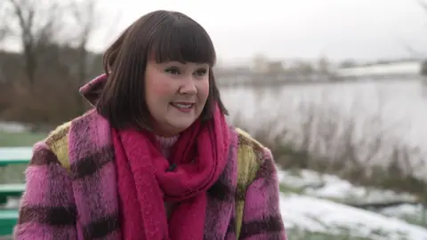 A woman with brown short hair and a full fringe, wears a pink scarf and pink checked coat, looking off to the camera talking, as she sits outside on a wintery day